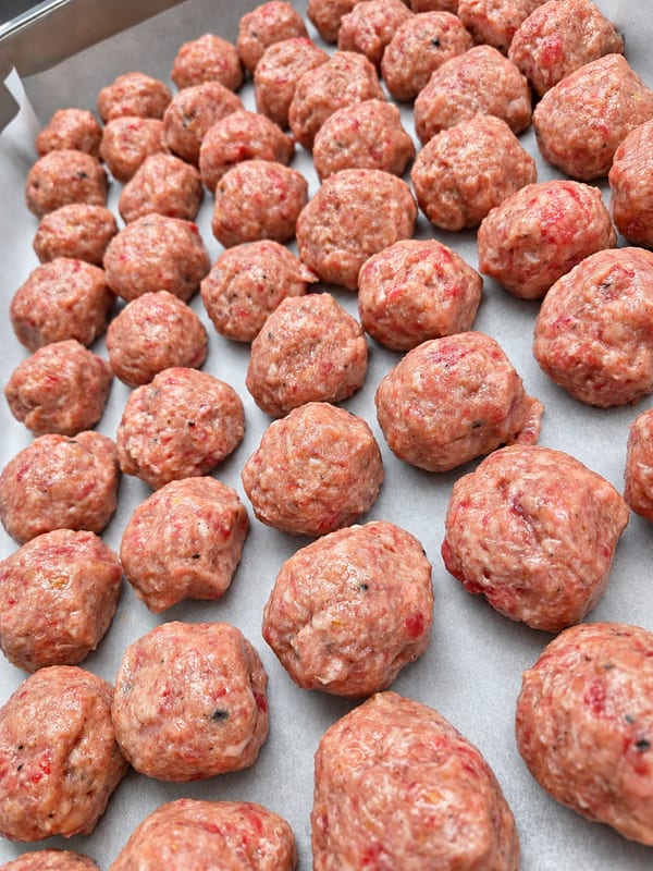 Handrullade råa köttbullar på en plåt med bakplåtspapper, före tillagning.